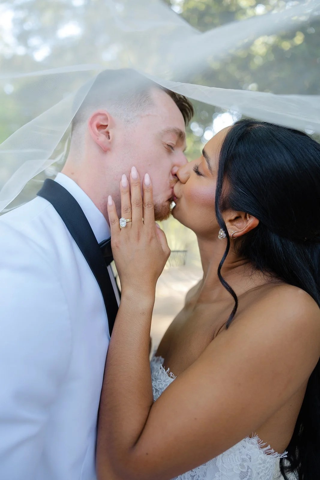 A bride and groom kiss under a sheer veil, with a close-up view of their faces and the bride's hand showing an engagement ring. The groom is dressed in a tuxedo, and the bride has long dark hair and wears earrings.