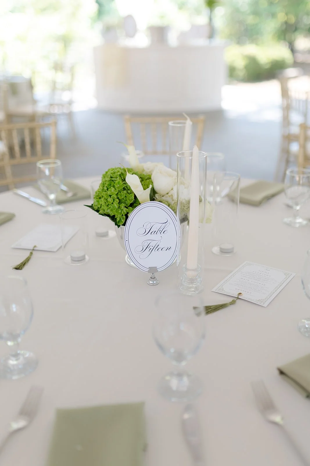 Elegant wedding reception table setup with white tablecloth, green floral centerpiece, a sign indicating table fifteen, tall white candles, clear glassware, and place cards with tassels.