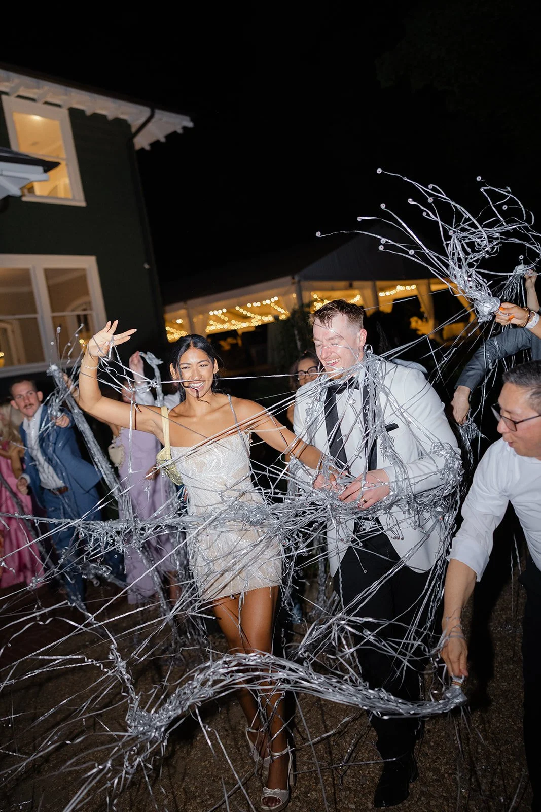 People celebrating at a nighttime wedding reception with silver streamers and strings of lights in the background.