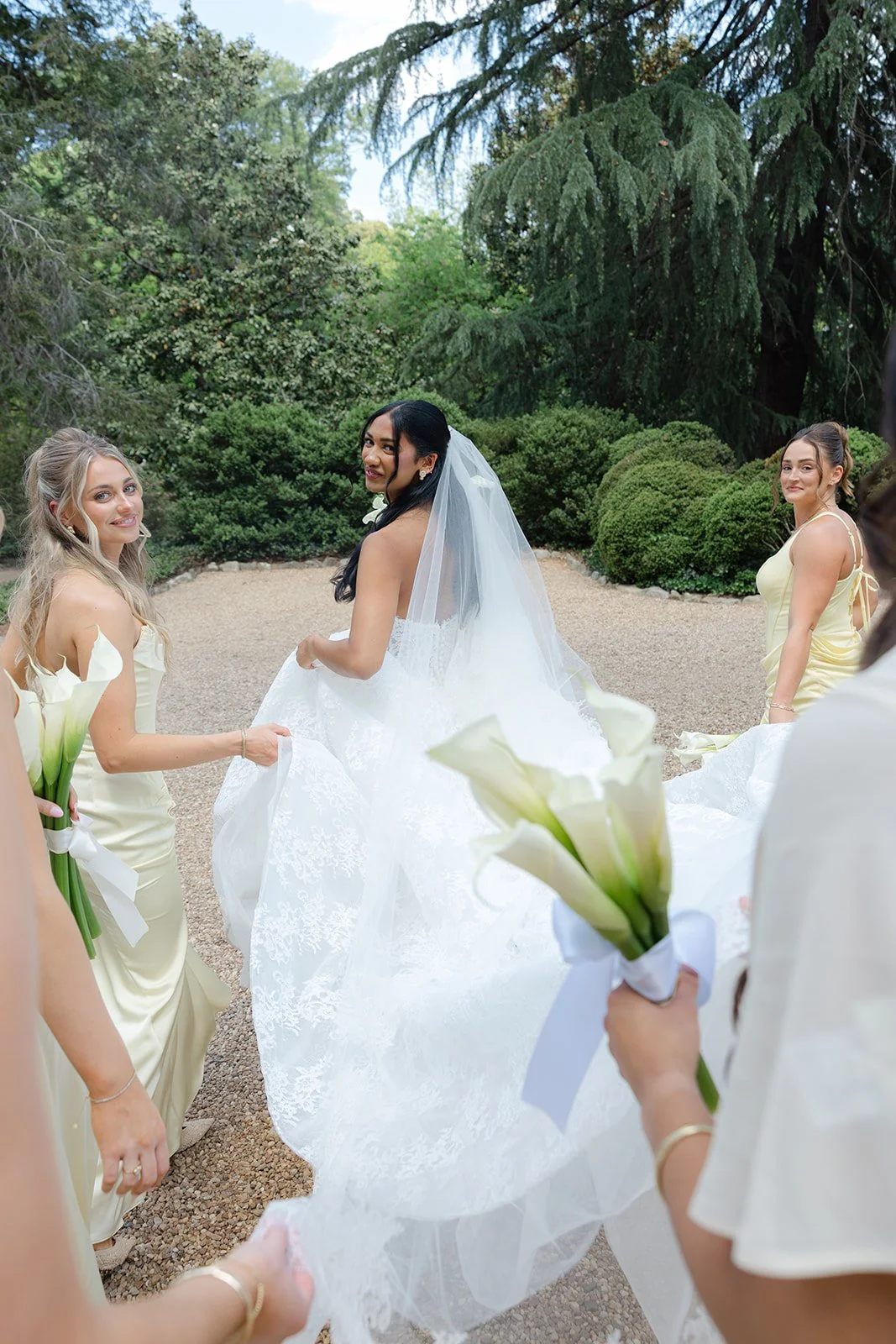 A bride in a wedding gown and veil holding her dress, surrounded by bridesmaids in yellow dresses holding calla lily bouquets, in a lush outdoor garden setting.