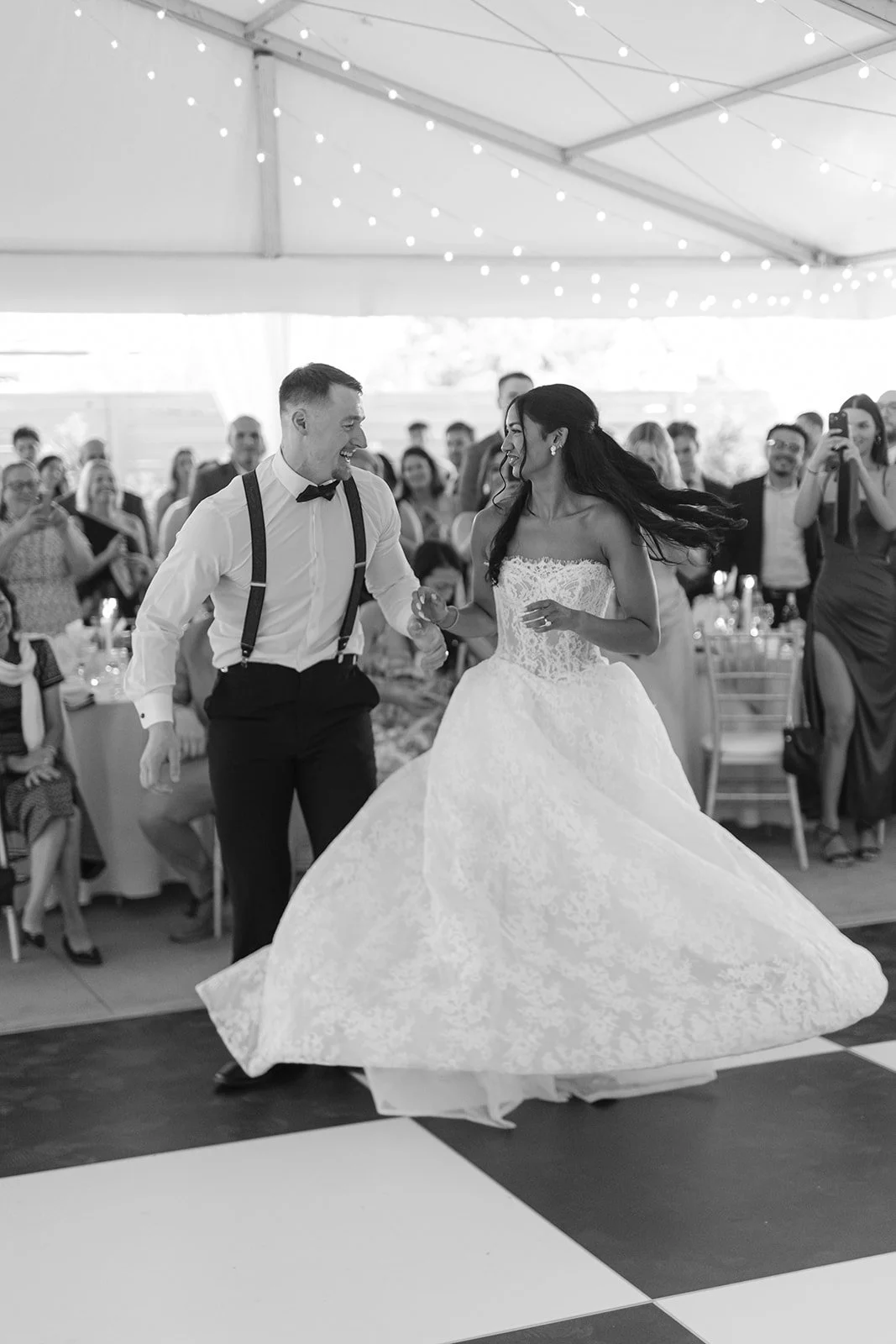 A black and white photo of a bride and groom dancing at their wedding reception, surrounded by guests seated and standing, under string lights.