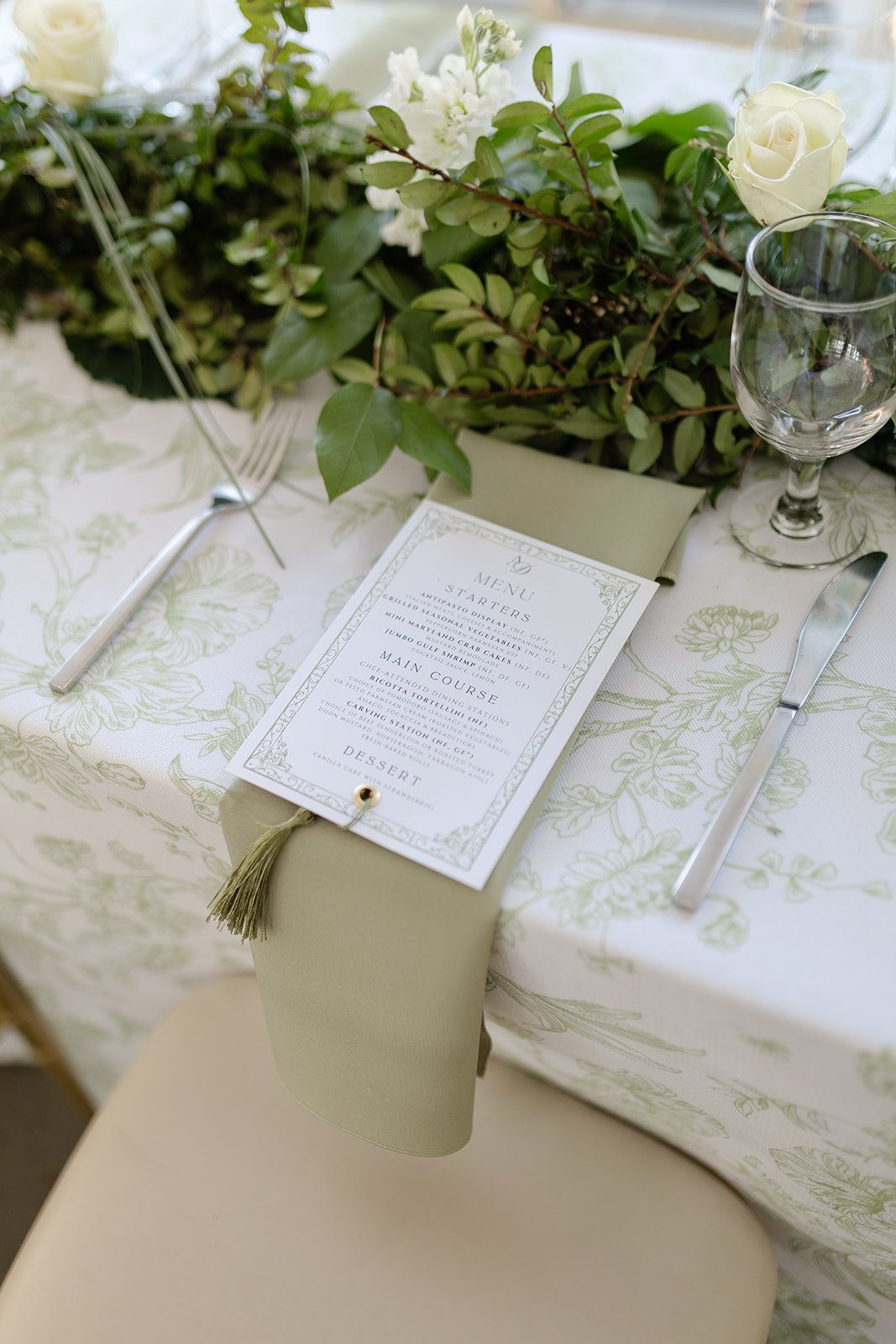 Elegant table setting with a menu card, cutlery, a glass, and a floral centerpiece with white roses and greenery.