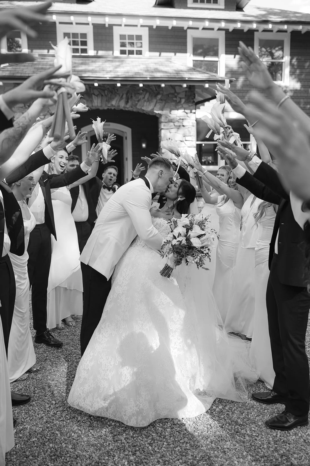 A black and white photo of a bride and groom kissing under a canopy of friends and family holding and throwing flowers, celebrating their wedding.