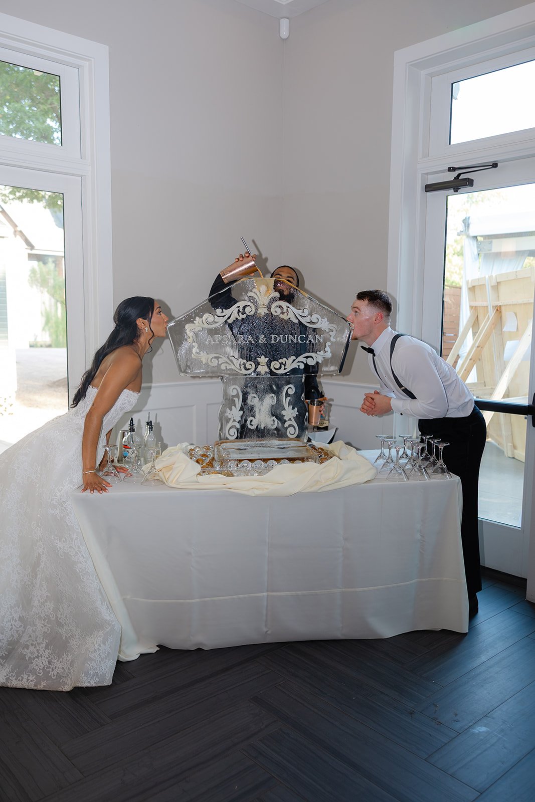 Bride and groom at wedding reception enjoying ice sculpture toast with champagne, standing on either side of ice sculpture with their names, 'Apsara & Duncan' engraved, inside a bright room with large windows, drinking glasses on table.