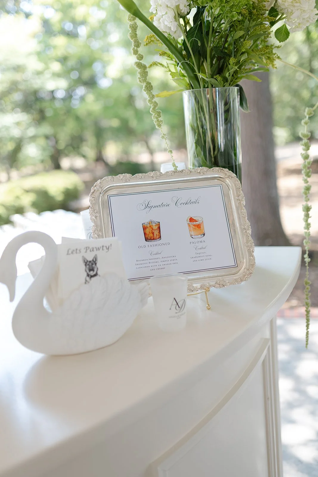 Elegant table setup with a large glass vase filled with white and green flowers, a decorative sign listing signature cocktails, a cup with a dog illustration and the phrase 'Let's Pawty!', and a small white container with initials.