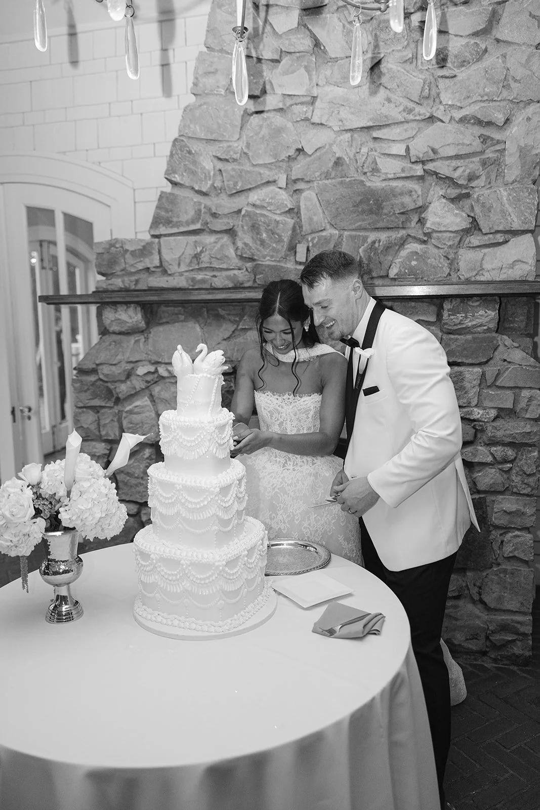 A bride and groom cutting a wedding cake at their reception, with the bride in a lace wedding dress and the groom in a tuxedo, standing in front of a stone wall, smiling.