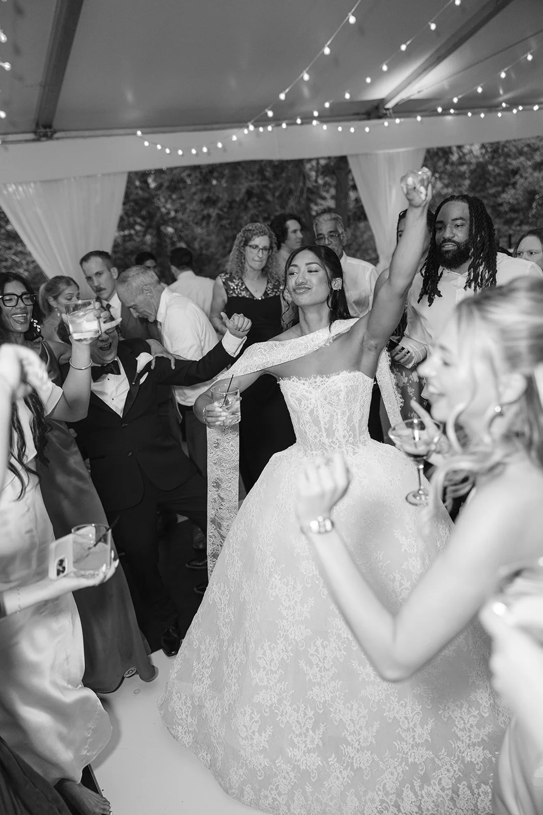 Black and white photo of a wedding reception with a bride in a lace gown dancing with guests, others holding drinks and celebrating under a canopy decorated with string lights.