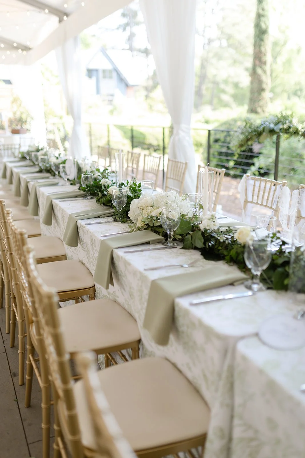 Elegant outdoor wedding reception table decorated with white and green floral arrangements, set with tableware and surrounded by beige chairs under a white canopy.