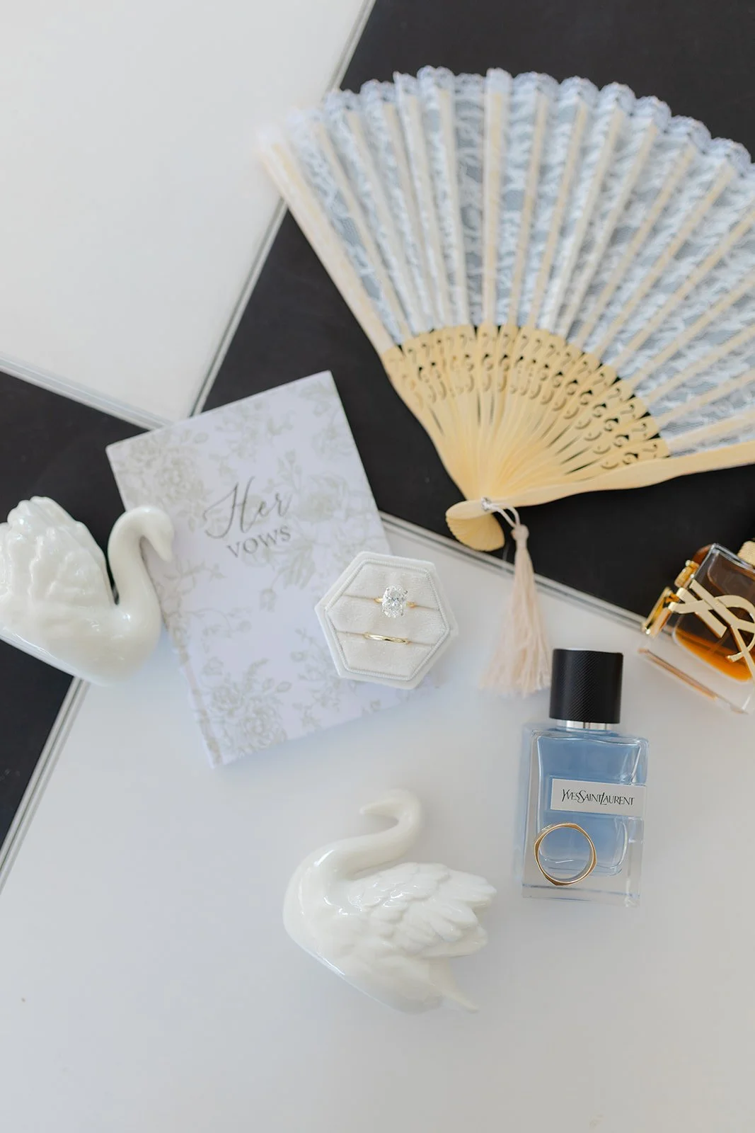 A collection of wedding or engagement items including a white swan porcelain figurine, a wedding vows card, a ring box with rings, a bottle of Yves Saint Laurent perfume, a floral hand fan, and decorative swan figures, arranged on a white surface.