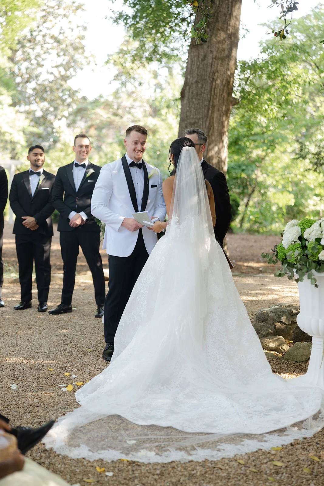 A wedding ceremony outdoors with the bride and groom exchanging vows, surrounded by groomsmen in black tuxedos and a large tree, with sunlight filtering through the leaves.