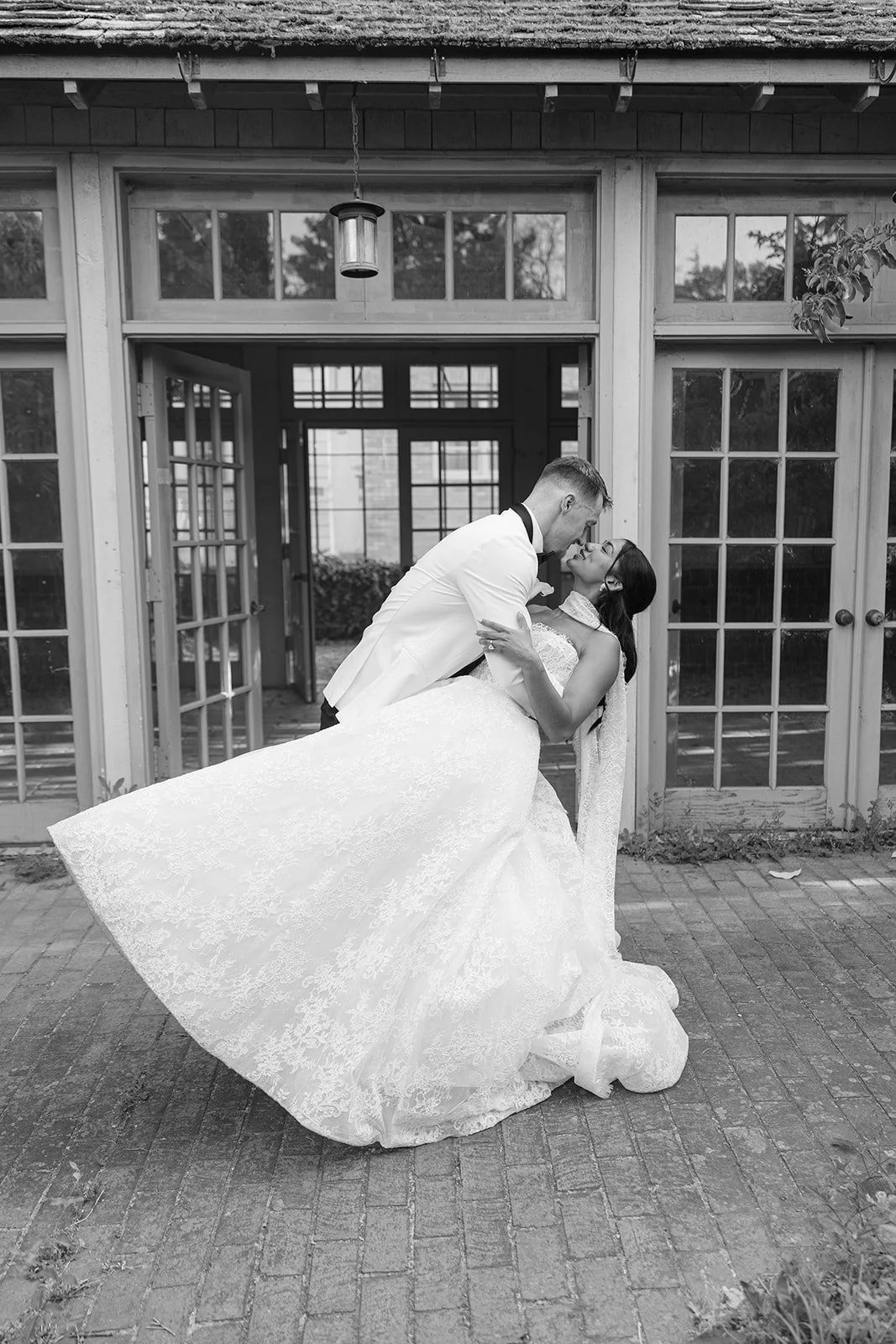 A newlywed couple sharing a romantic dip outside a building with glass doors and windows, black and white photo.