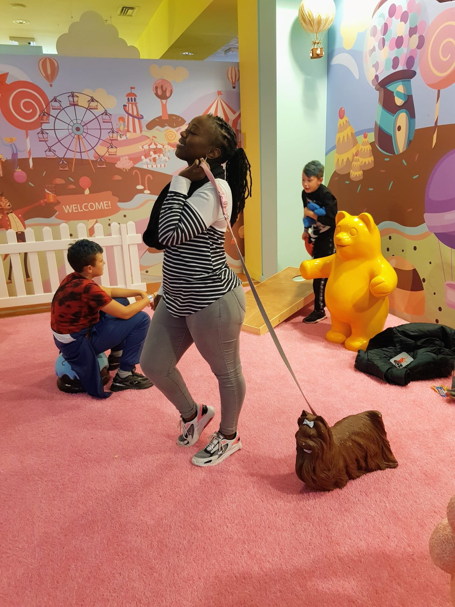 A colorful indoor play area with cartoon-style murals, a yellow bear-shaped sculpture, Children playing and an adult woman smiling with a dog on a leash. The background features a carnival scene with a Ferris wheel, carousel, and balloons.