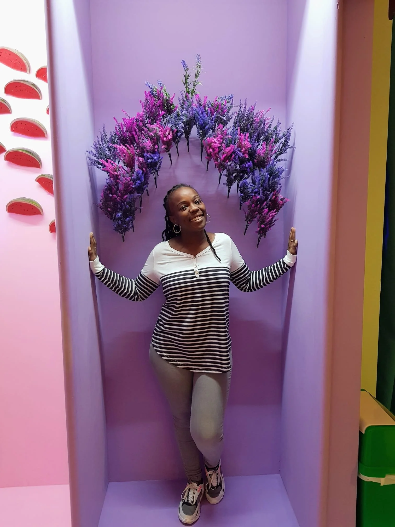 Woman with braided hair smiling, wearing hoop earrings, a striped shirt, and beige pants, standing inside a purple cubicle decorated with purple and pink flowers.