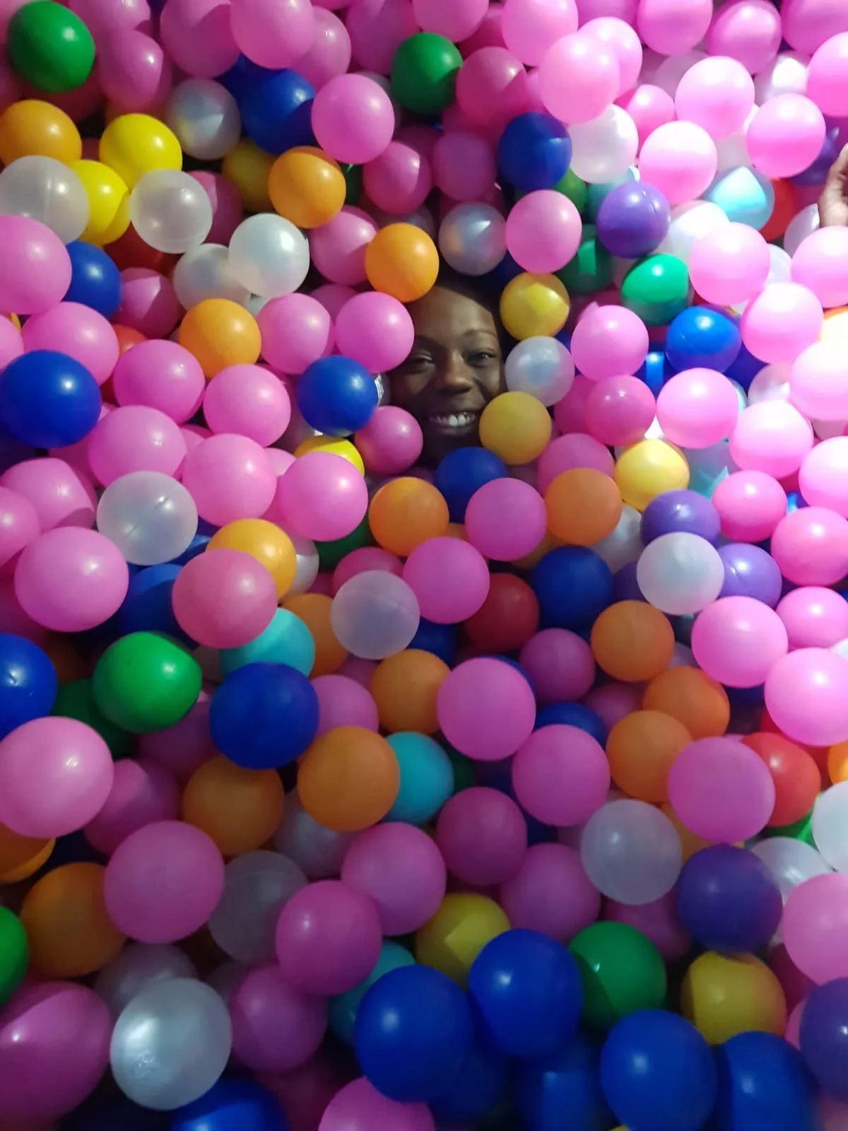 Person's face partially visible among colorful plastic balls at a ball pit, smiling with eyes closed.
