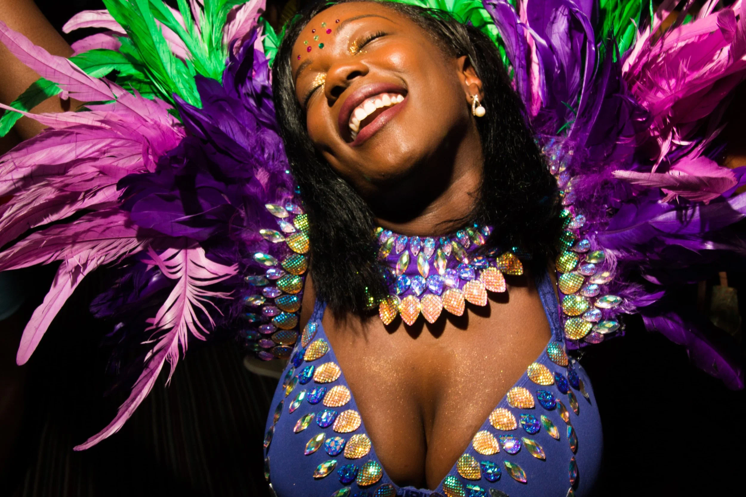 A smiling woman dressed in a vibrant purple and green carnival costume adorned with feathers and sparkling stones.