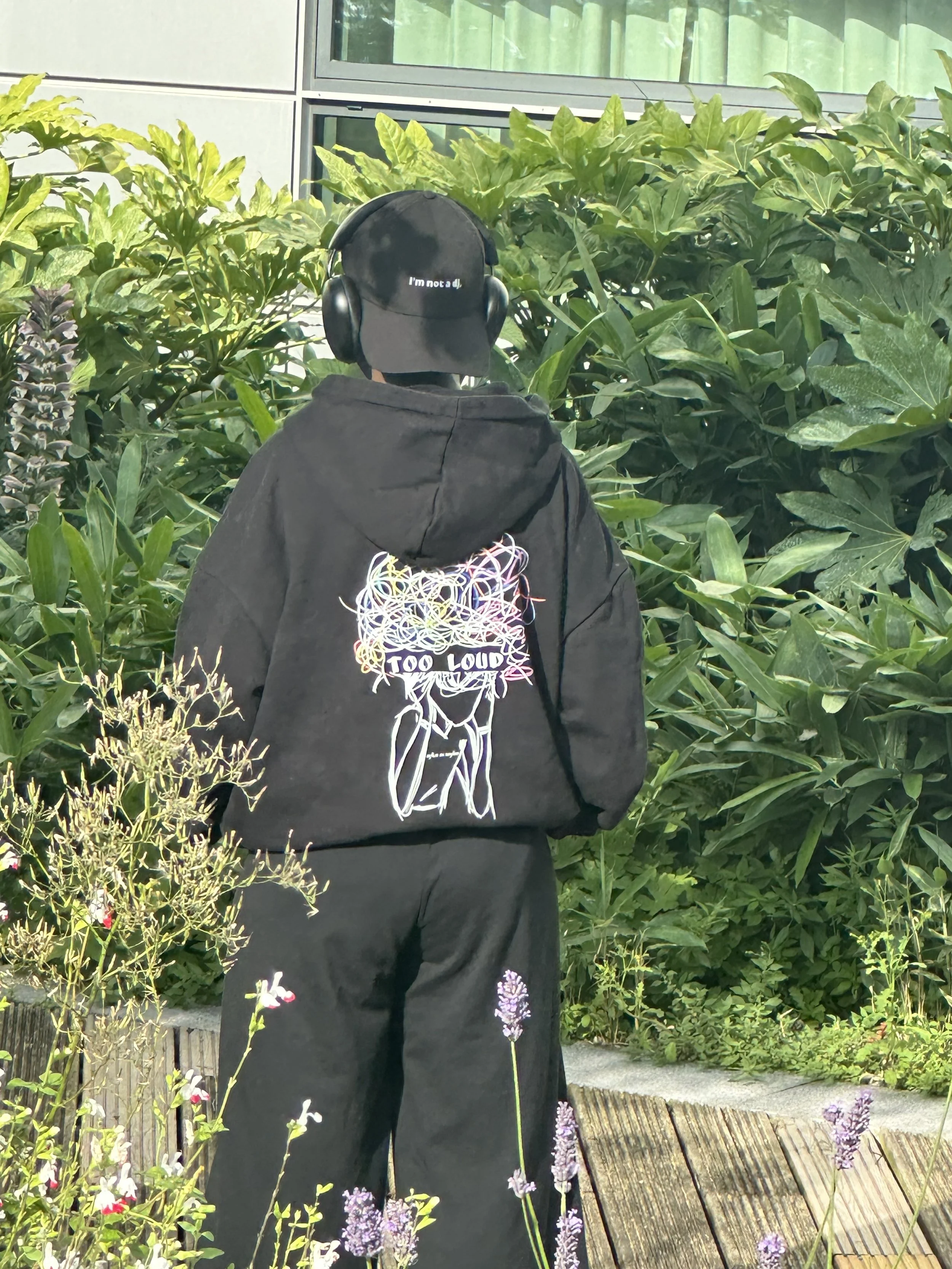 A person standing in front of green bushes, wearing a black hoodie with a colorful graphic and the text 'TOO LOUD', a black baseball cap with the words 'I'm not a DJ', and large black headphones.