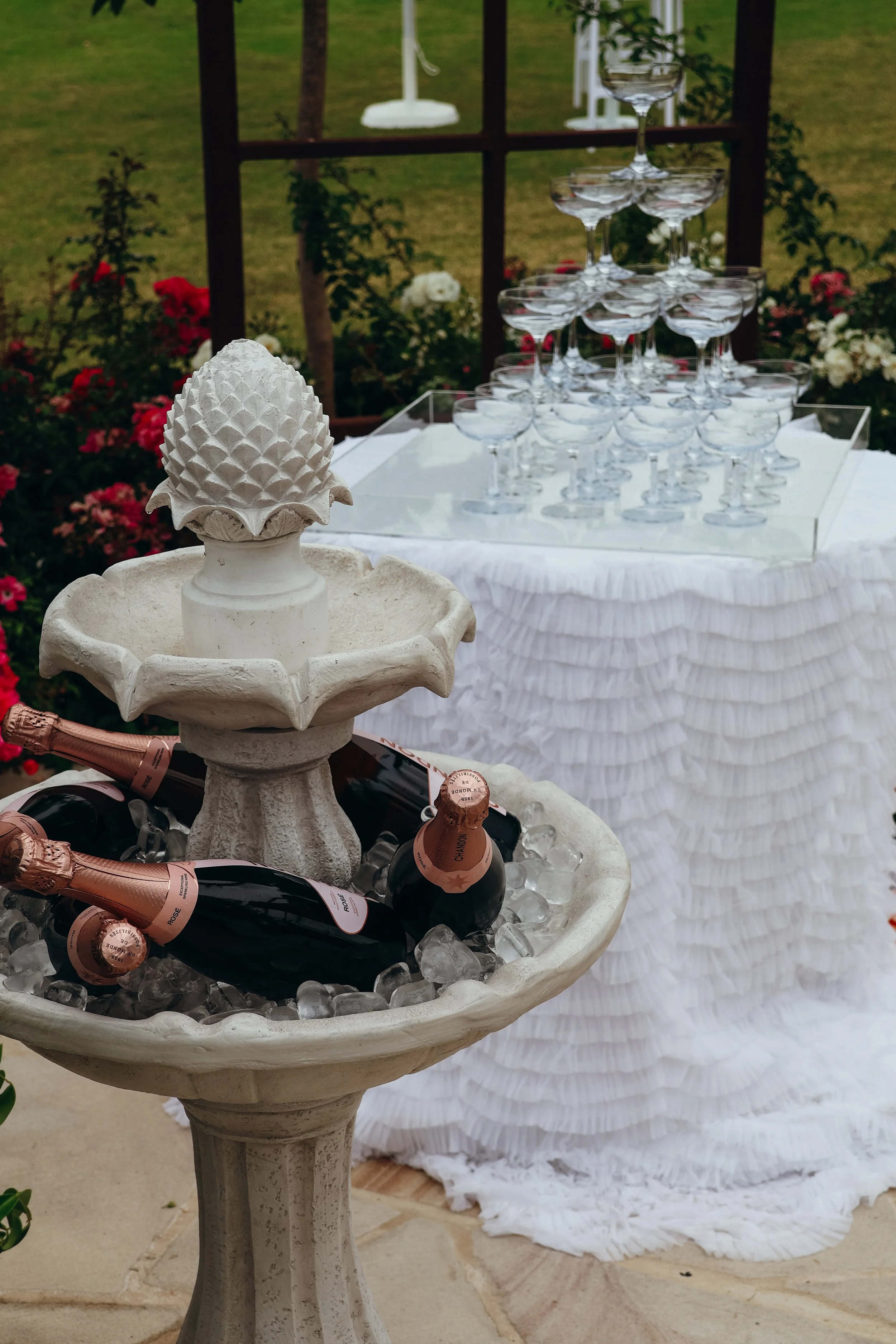 Champagne bottles in an ice bucket in front of a tiered Cinderella glass pyramid at an outdoor event, with a white ruffled tablecloth and flowers in the background.