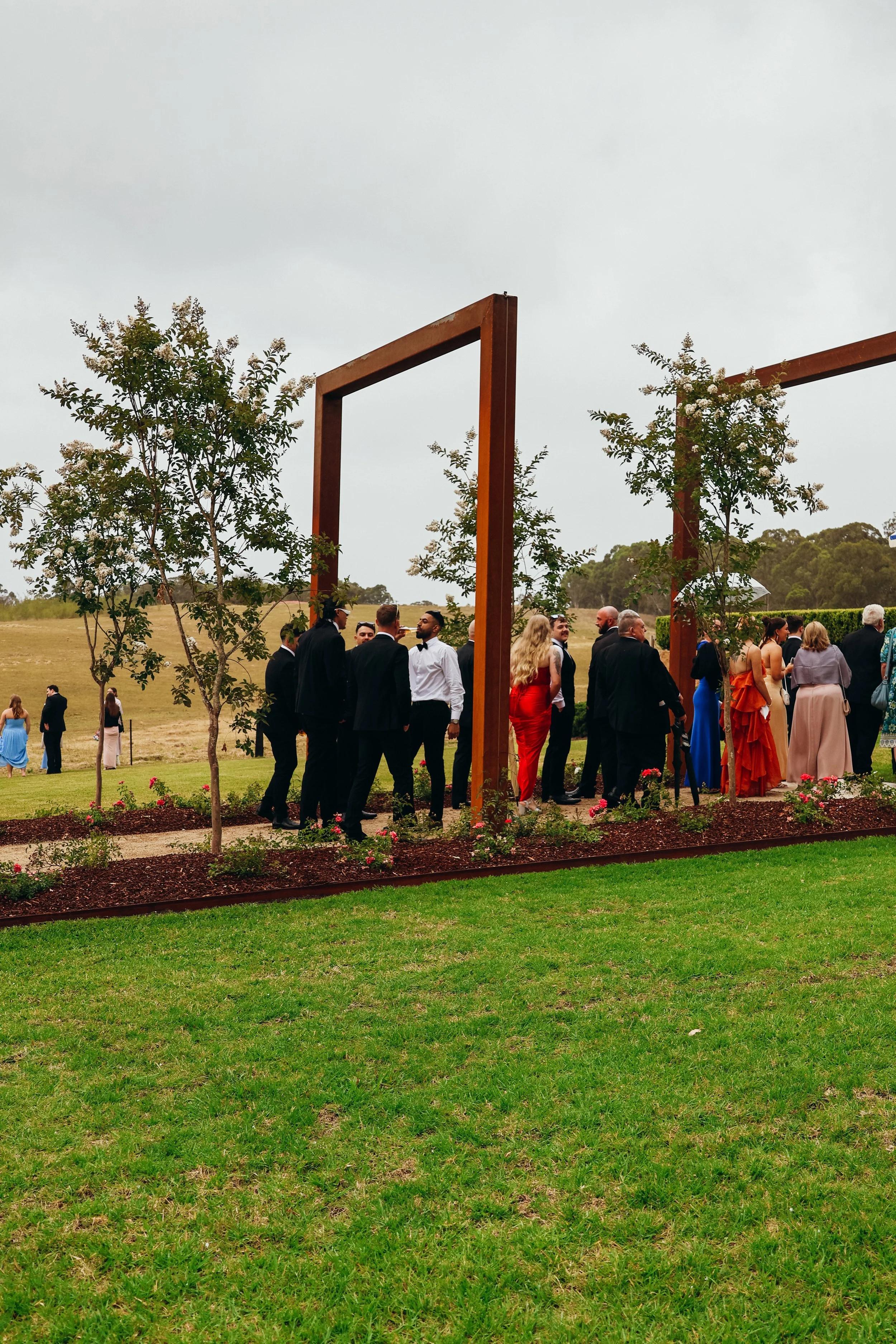 People dressed in formal attire at an outdoor event under a modern wooden archway, with trees and a grassy field in the background.