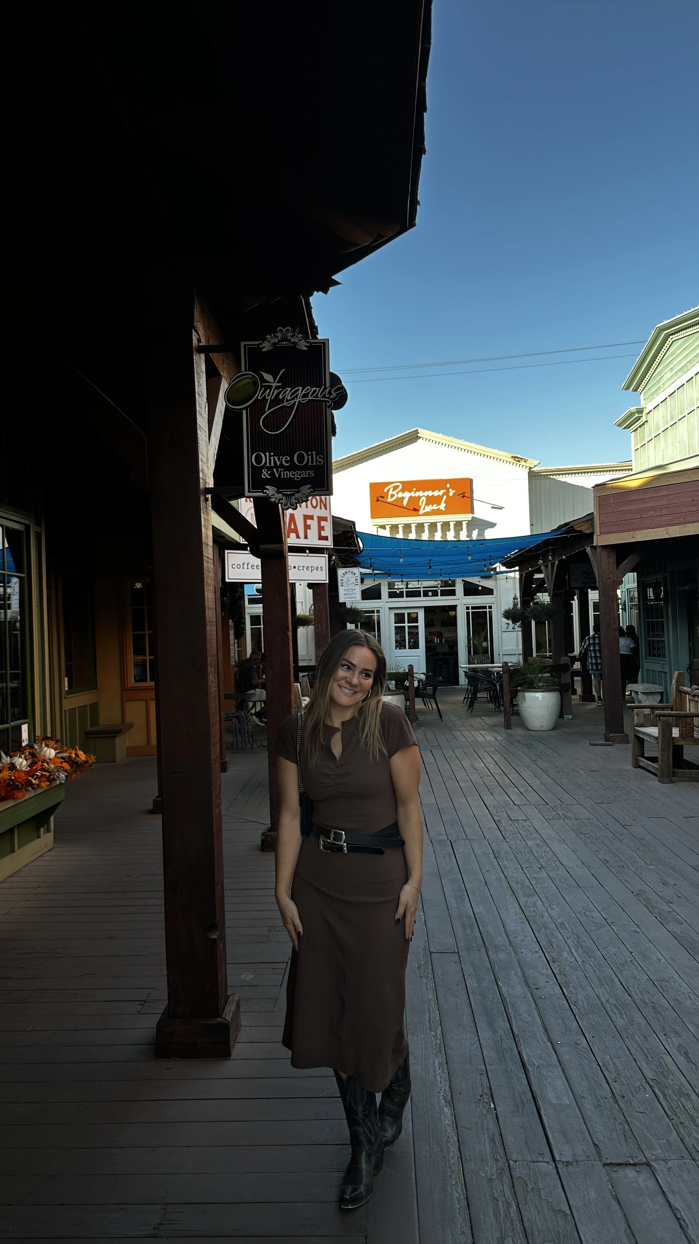 A young woman with long hair standing on a wooden deck in a shopping or dining area, smiling and wearing a brown dress with a black belt and cowboy boots, with shops and signs in the background.