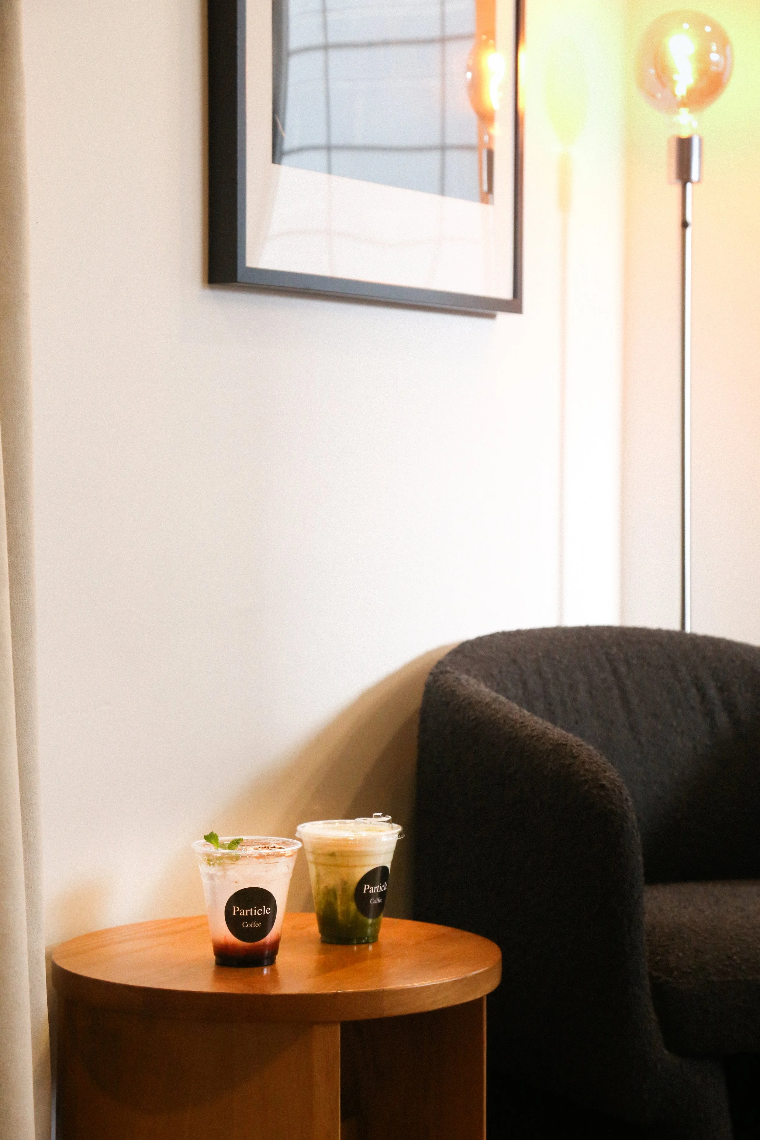 Two cups of iced coffee on a round wooden table beside a dark armchair, with a framed picture and a floor lamp in the background.