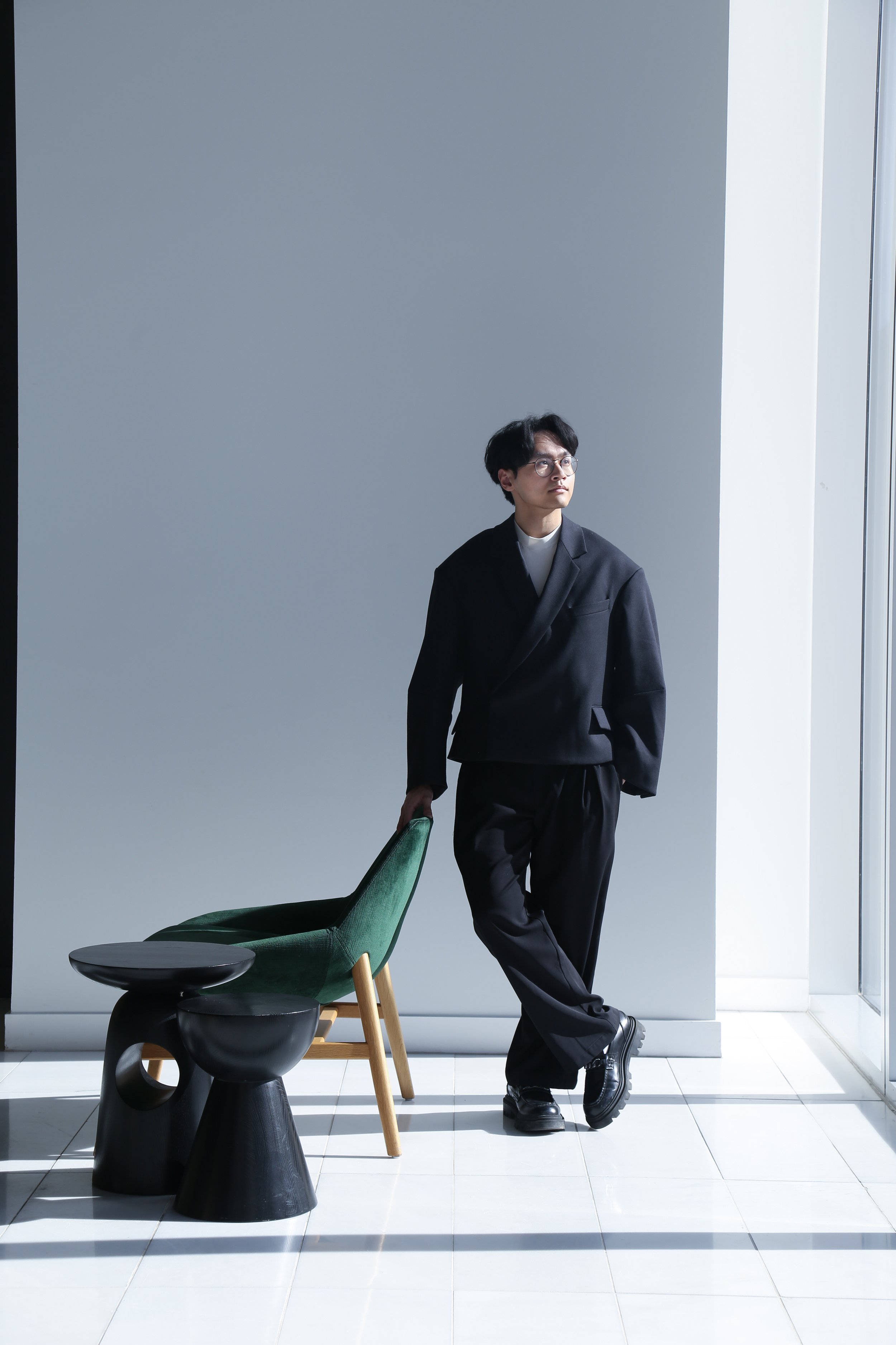 A man in black formal attire with glasses, standing in front of a white wall and large window, holding a green chair.