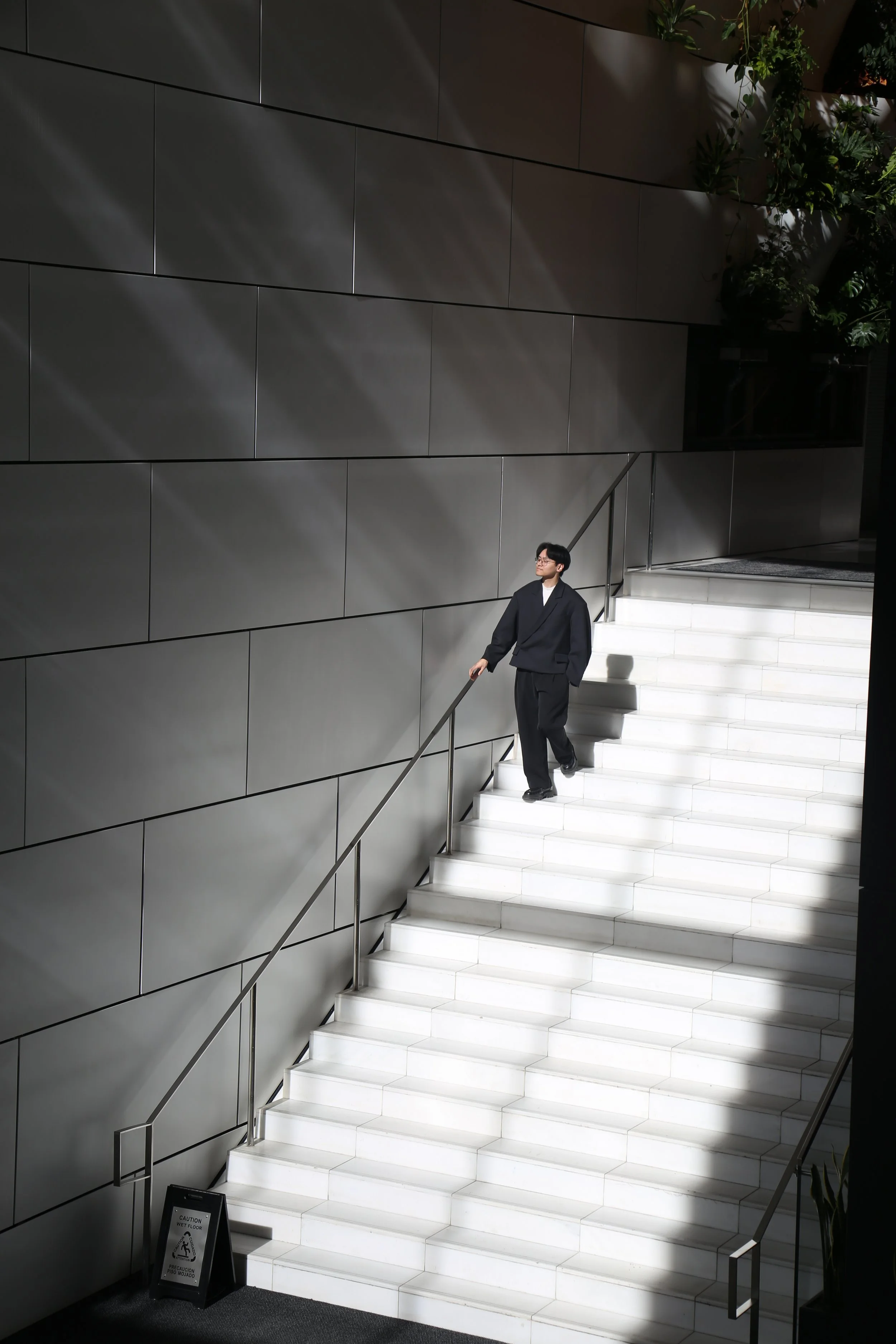 A person dressed in dark clothing climbing white marble stairs in a modern indoor setting, with a gray tiled wall and some greenery in the background.
