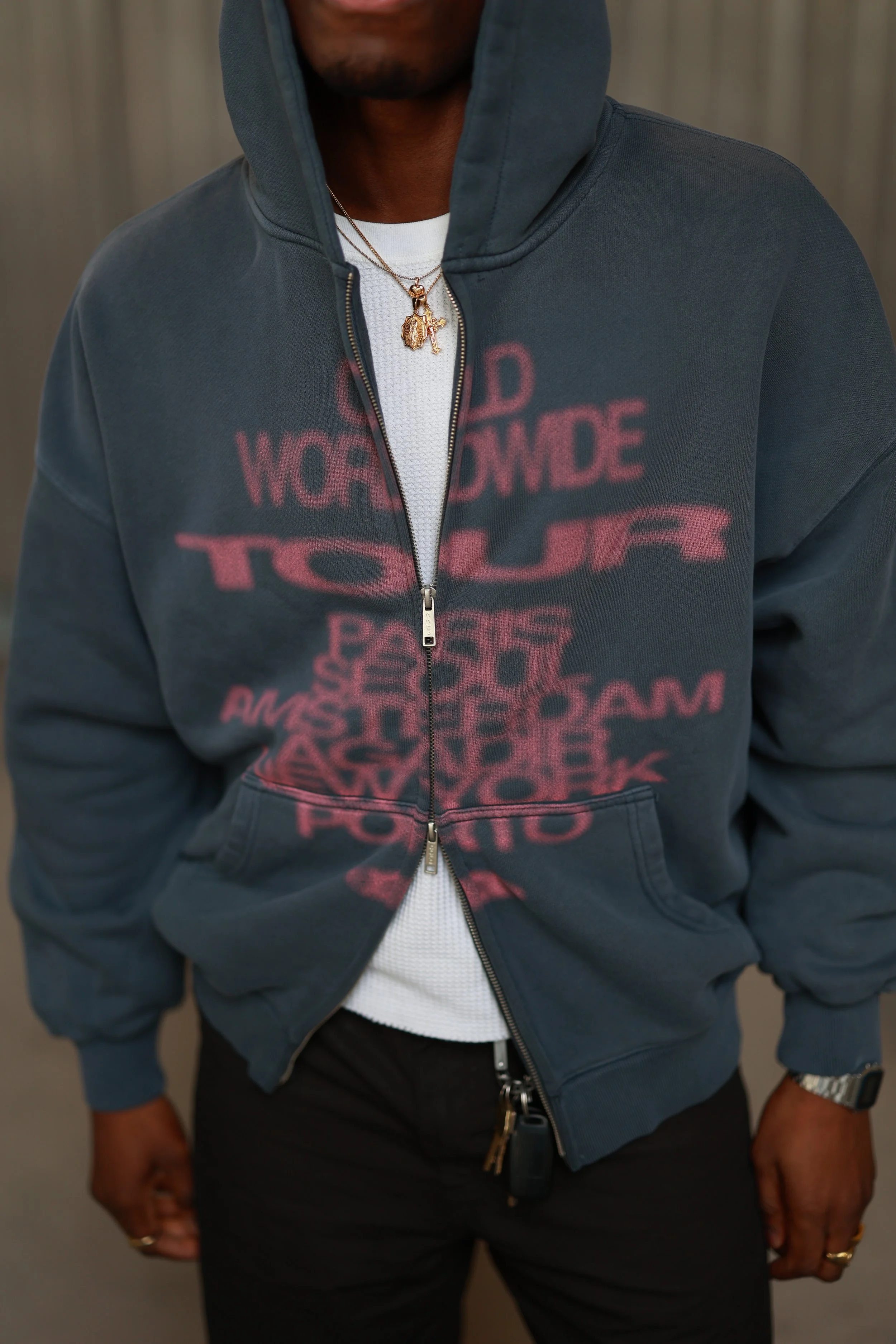 Person wearing a dark blue hooded jacket with red text and graphics, a white shirt underneath, black pants, gold jewelry including a necklace and rings, and a watch.