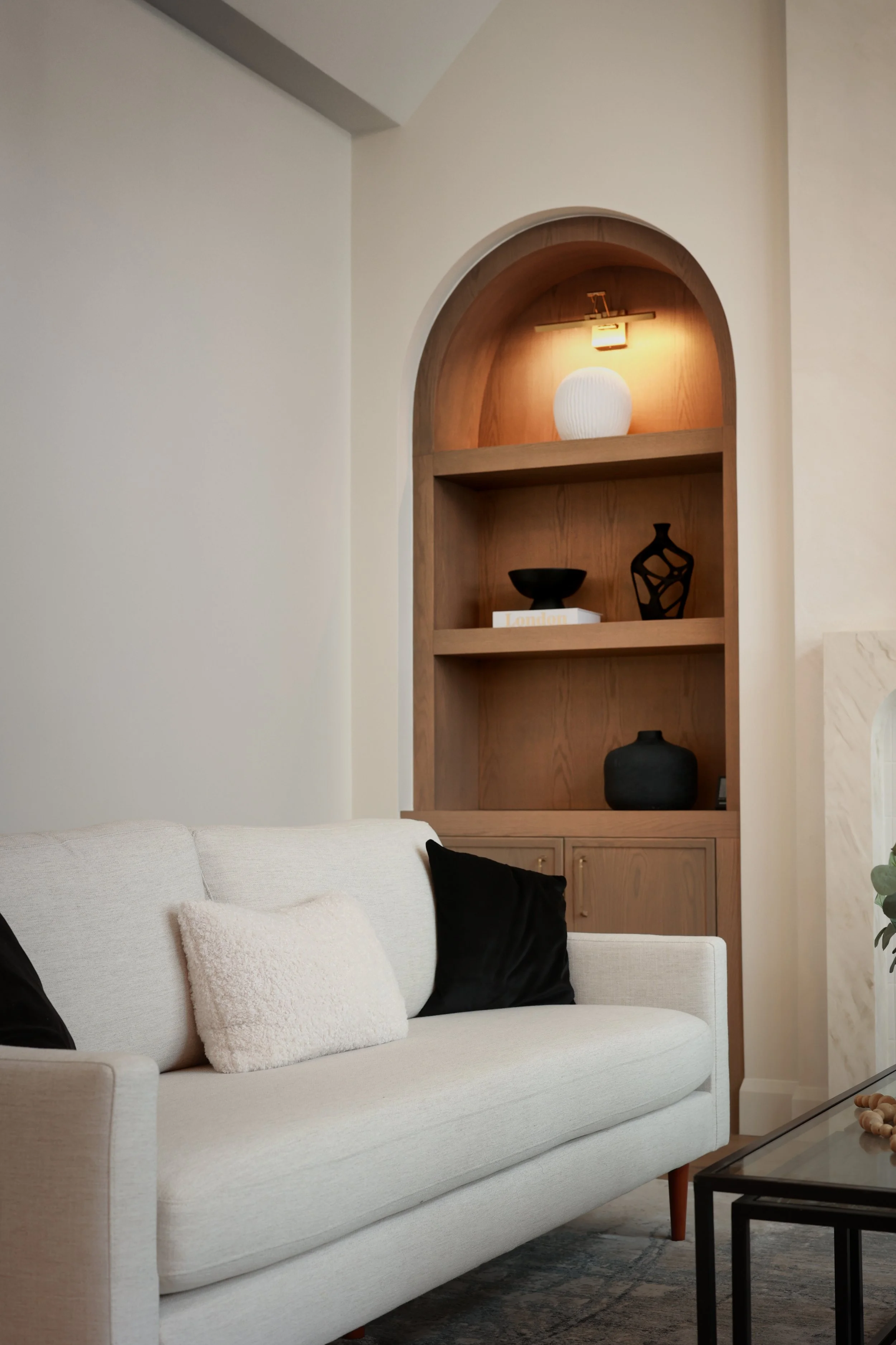 Living room with a white sofa, black and white pillows, and a built-in wooden shelf with decorative items, illuminated by a wall light.