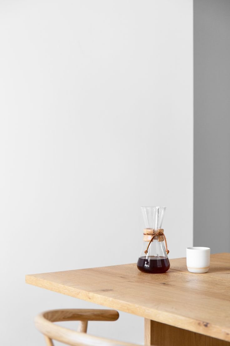 A wooden table with a glass coffee decanter filled with dark coffee, tied with a brown ribbon, and a small white cup on a plain white wall background.