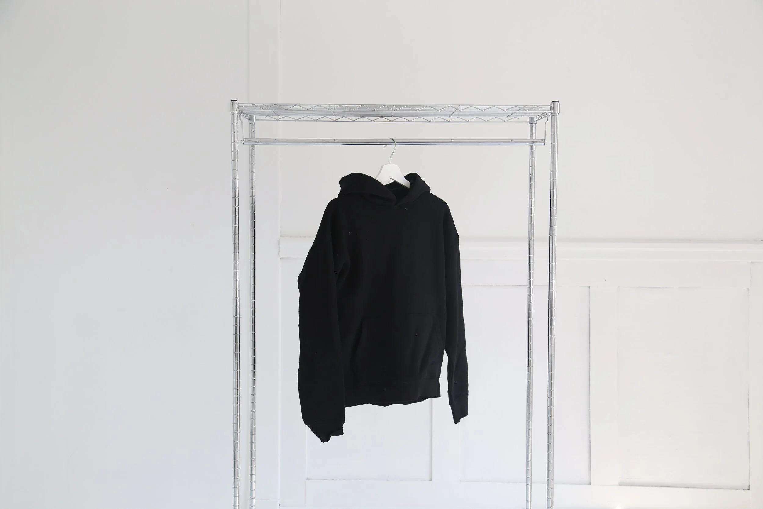 A black hoodie hanging on a white hanger from a metal clothing rack against a white wall.