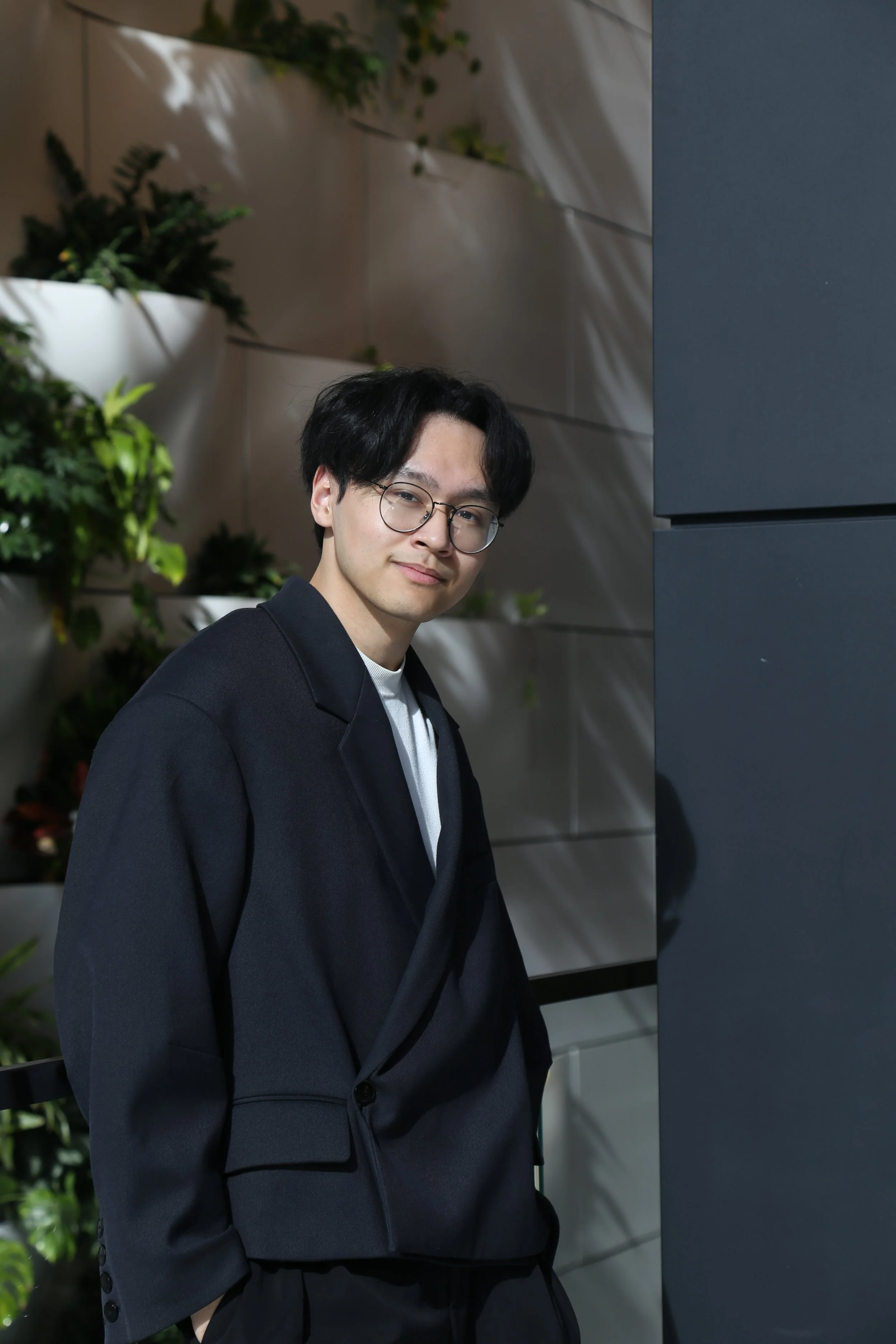 A young man with black hair and glasses, wearing a black blazer and white shirt, standing next to a dark wall with green plants in the background.