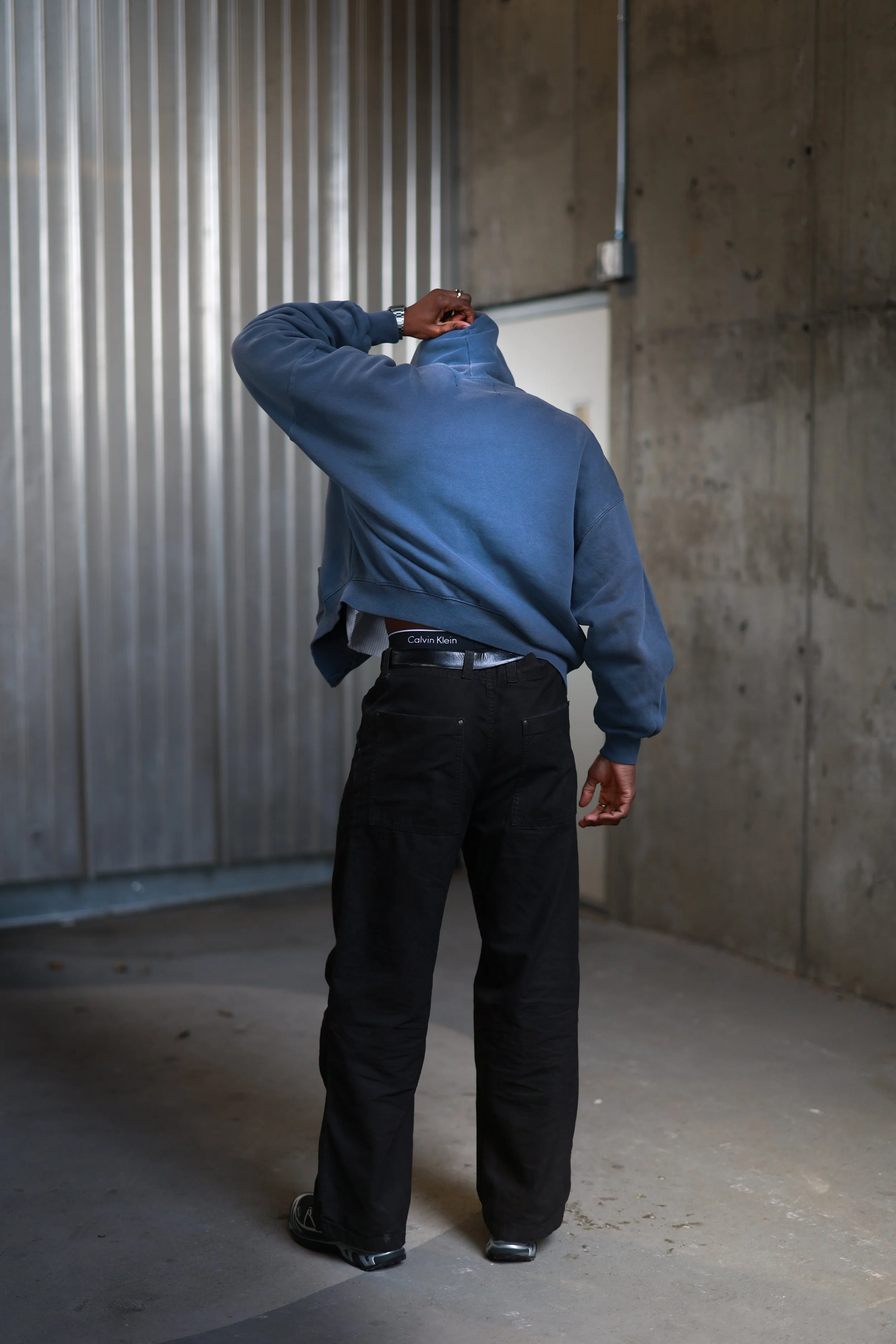 Person standing in an industrial space with concrete walls, wearing a blue hoodie, black pants, and Calvin Klein underwear, adjusting the hoodie.