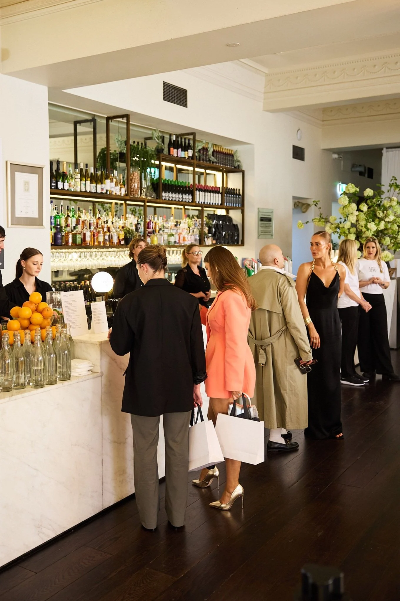 People standing at a bar in a stylish indoor venue with bottles and glasses displayed on shelves.