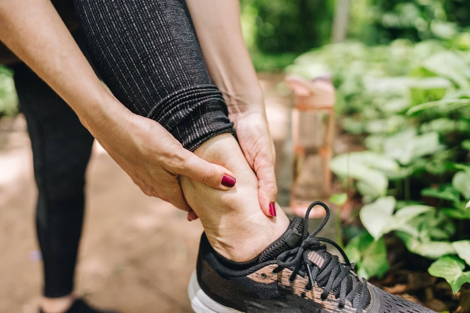 Person tying their ankle while wearing a black athletic shoe, in a green outdoor setting.
