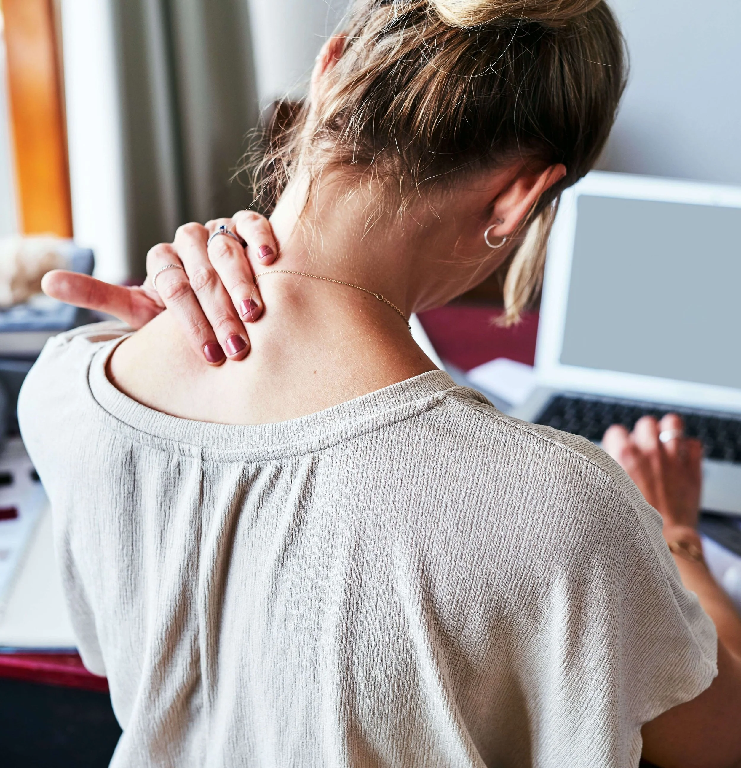 A woman with short hair, wearing a beige shirt, is holding her neck with her left hand while working on a laptop. She appears to be experiencing neck pain or discomfort.