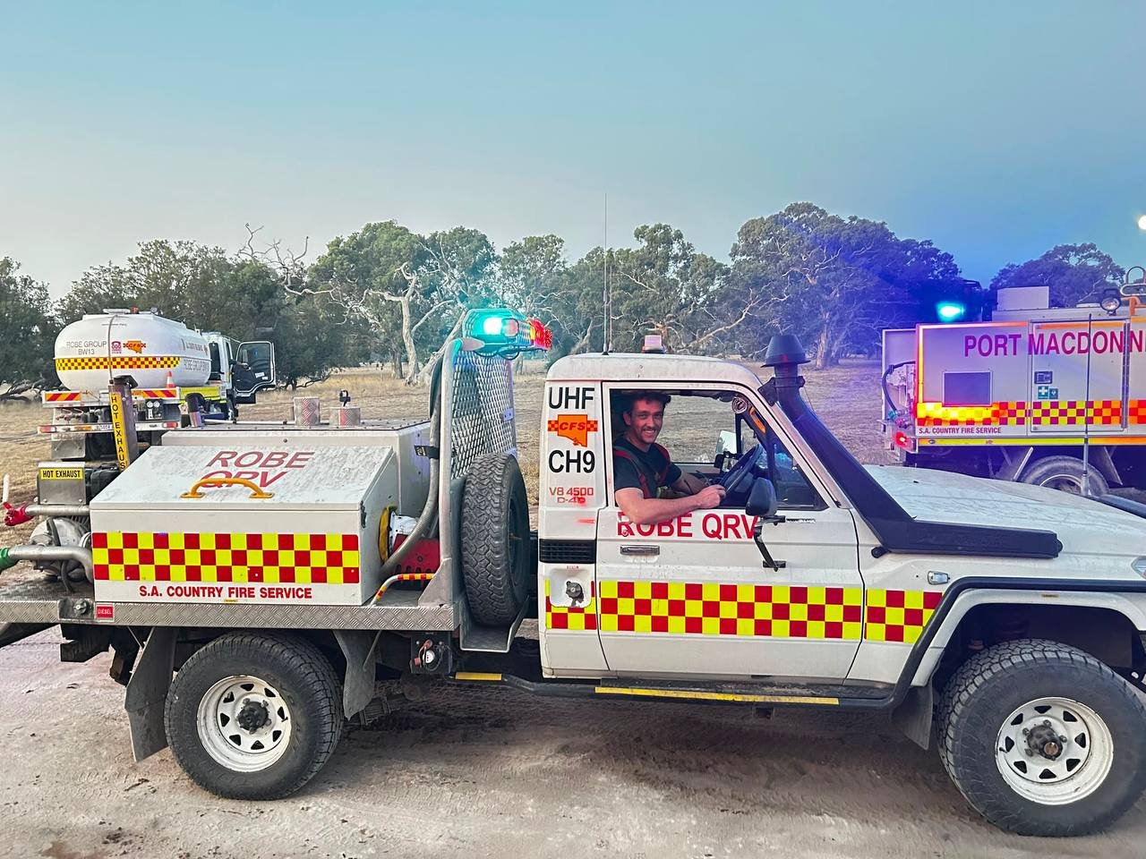 Firefighting vehicles from S.A. Country Fire Service parked outdoors with trees in the background; one vehicle has a smiling firefighter in the driver's seat.