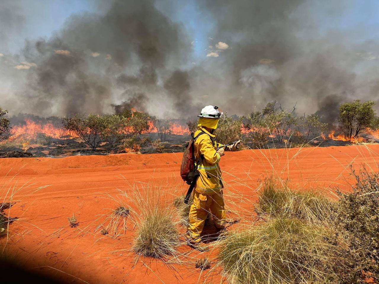 Firefighter in yellow protective gear and helmet fighting a wildfire in a desert landscape with orange sand, sparse vegetation, and large smoke plumes in the sky.