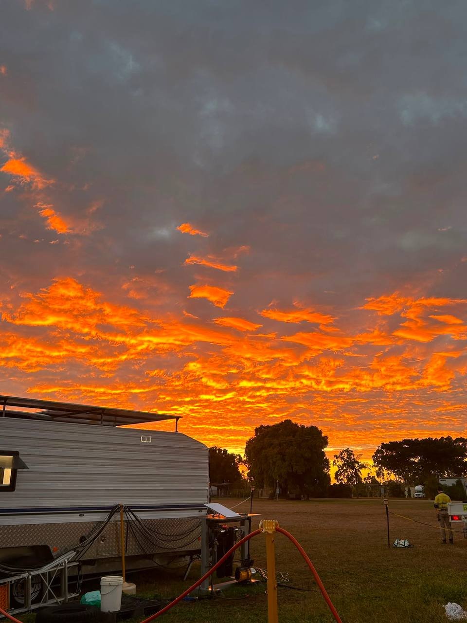 A vibrant orange and yellow sunset sky with dark gray clouds, viewed over an open grassy area with trees and some parked vehicles, including a camper trailer, and people in the distance.