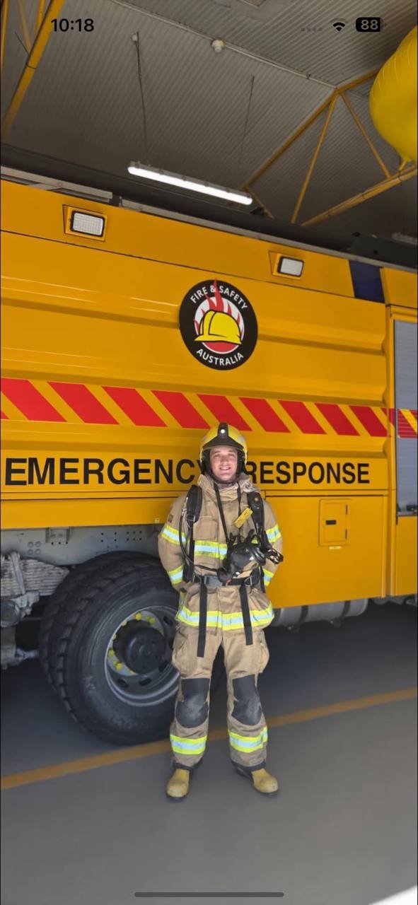 Firefighter standing in front of a yellow emergency response vehicle with fire safety Australia logo in a garage or station.