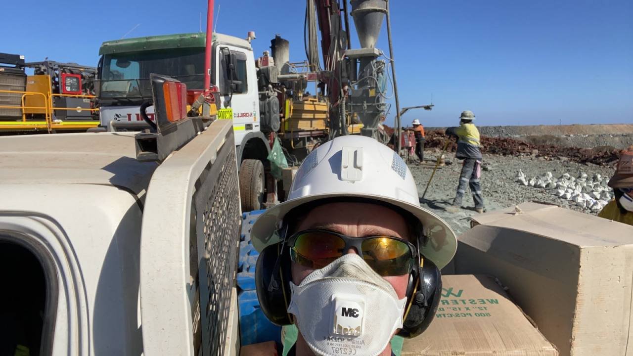 A person wearing a white hard hat, safety glasses, headphones, and a mask taking a selfie at a construction site with workers and heavy machinery in the background.