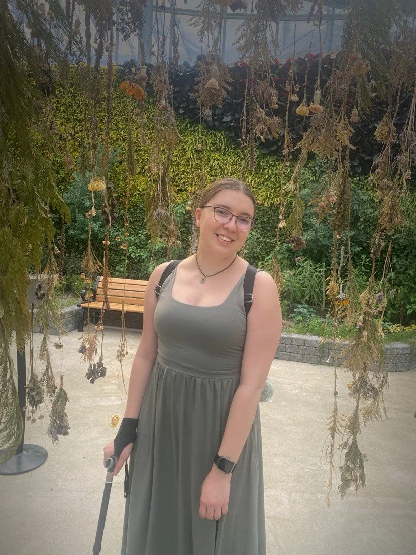 Alexus Wiebe stands in front of a greenery backdrop. she is wearing a light green dress and using forearm crutches.