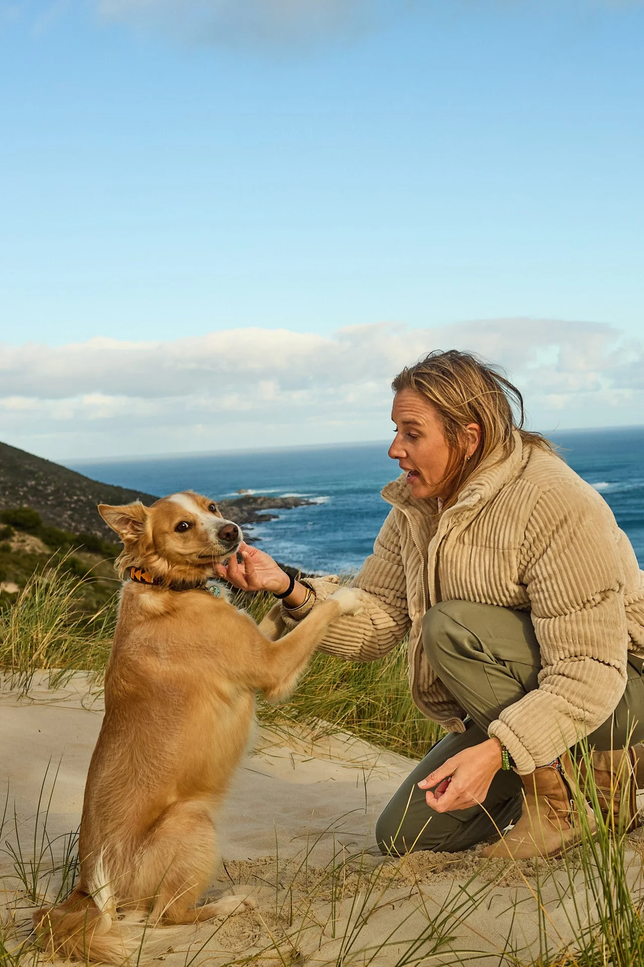 A woman in outdoor clothing kneeling on sandy dunes, holding hands with a dog against a backdrop of grass, hills, and an ocean with waves and clouds in the sky.