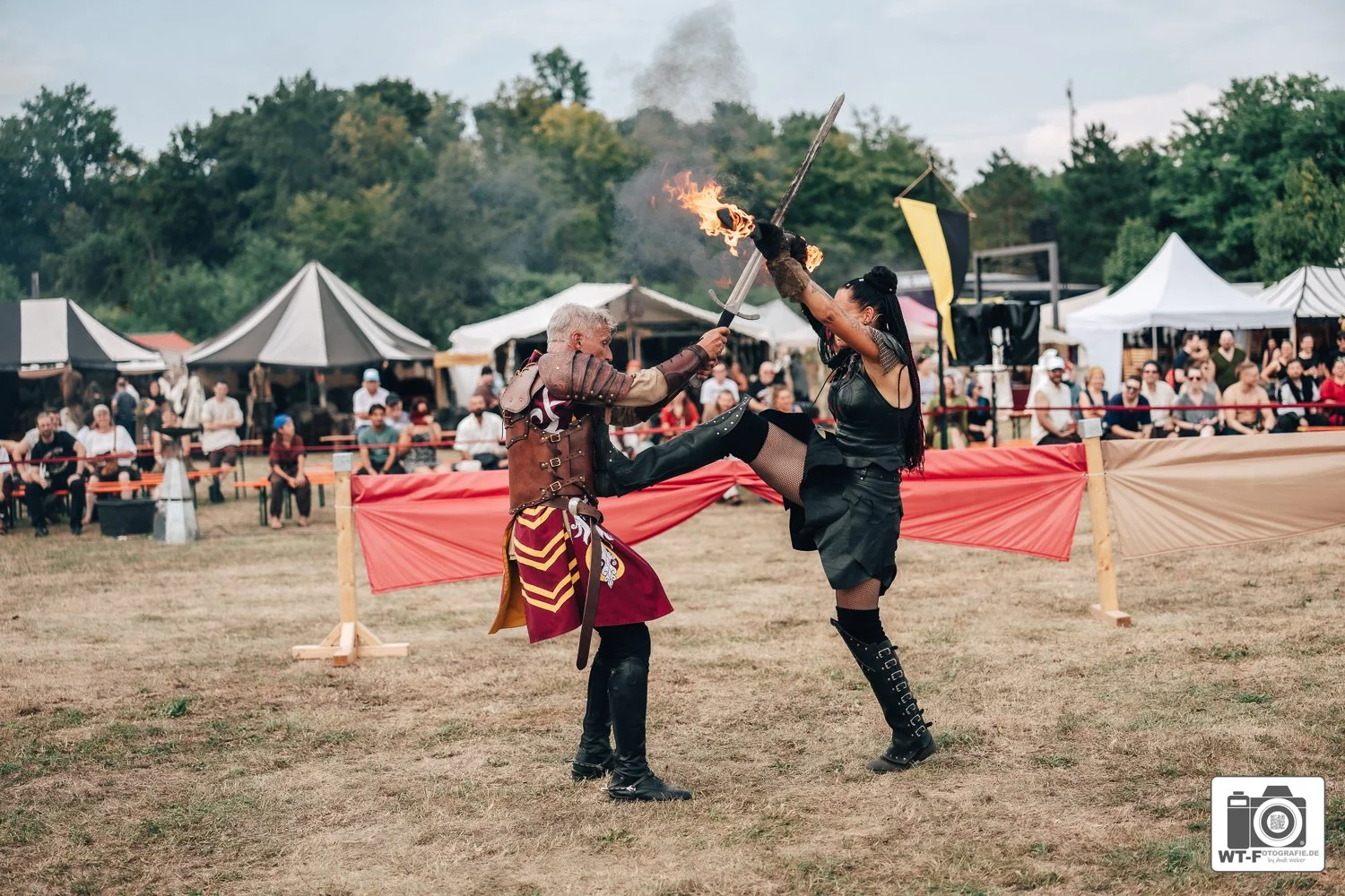 Zwei Menschen in mittelalterlicher Kleidung kämpfen mit Schwertern bei einem Ritterfest, während im Hintergrund ein Publikum zuschaut.