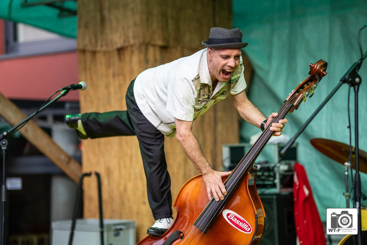 Ein Mann spielt auf einem Bass, während er ein Bein auf ein Bassinstrument stellt und laut schreit, auf einer Bühne mit Mikrofonen und Instrumenten.