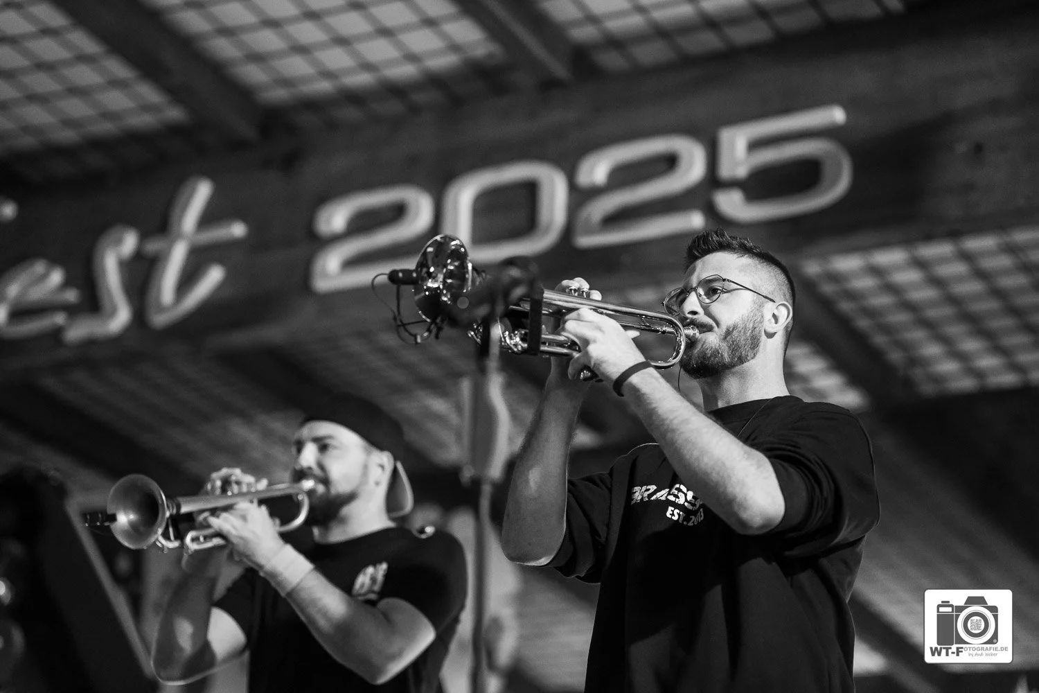 Zwei Männer Brass Buebe spielen Trompete in einem Raum mit Holzdecke und einer Wand, auf der 'est 2025' geschrieben steht. Das Bild ist schwarz-weiß.