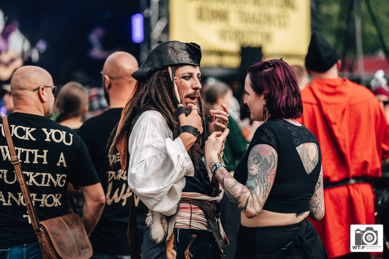 Zwei Frauen im Gespräch auf einem Open-Air-Konzert, eine trägt Piratenhut und ein Piratenkostüm, die andere hat Tattoos an den Armen. Im Hintergrund sind weitere Menschen, einige in roter Kleidung, und eine Bühne mit gelbem Banner sichtbar.