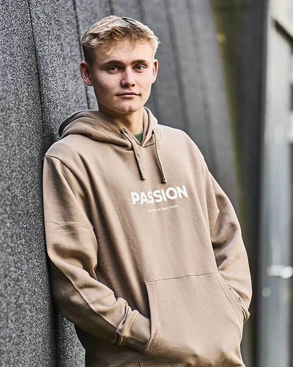 Ung mand i beige hoodie med teksten 'PASSION' står mod væg udendørs.