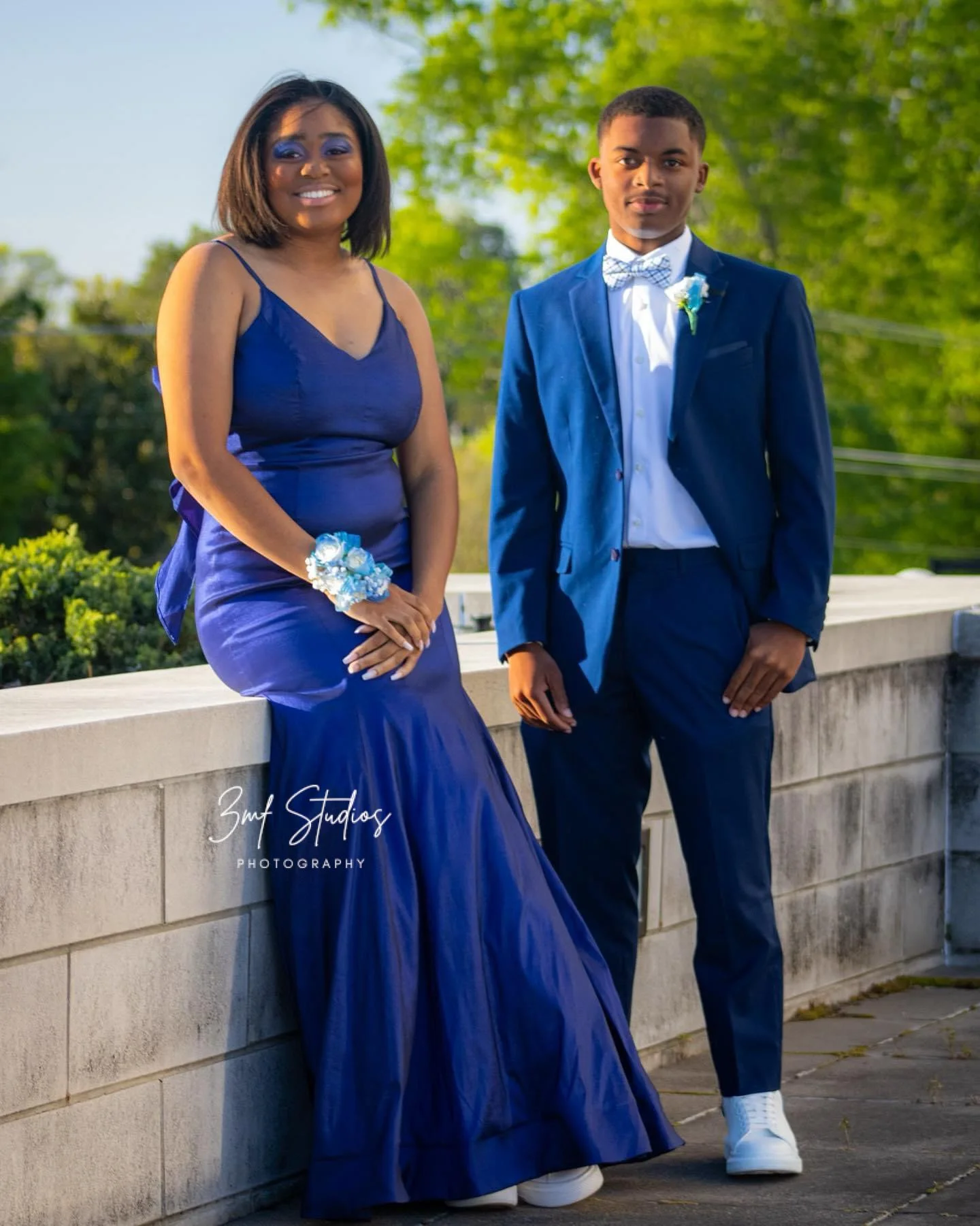 Young woman and young man dressed formally, standing outdoors by a stone wall with green trees in the background. The woman wears a long blue dress with a corsage on her wrist, and the man wears a blue suit with a bow tie.