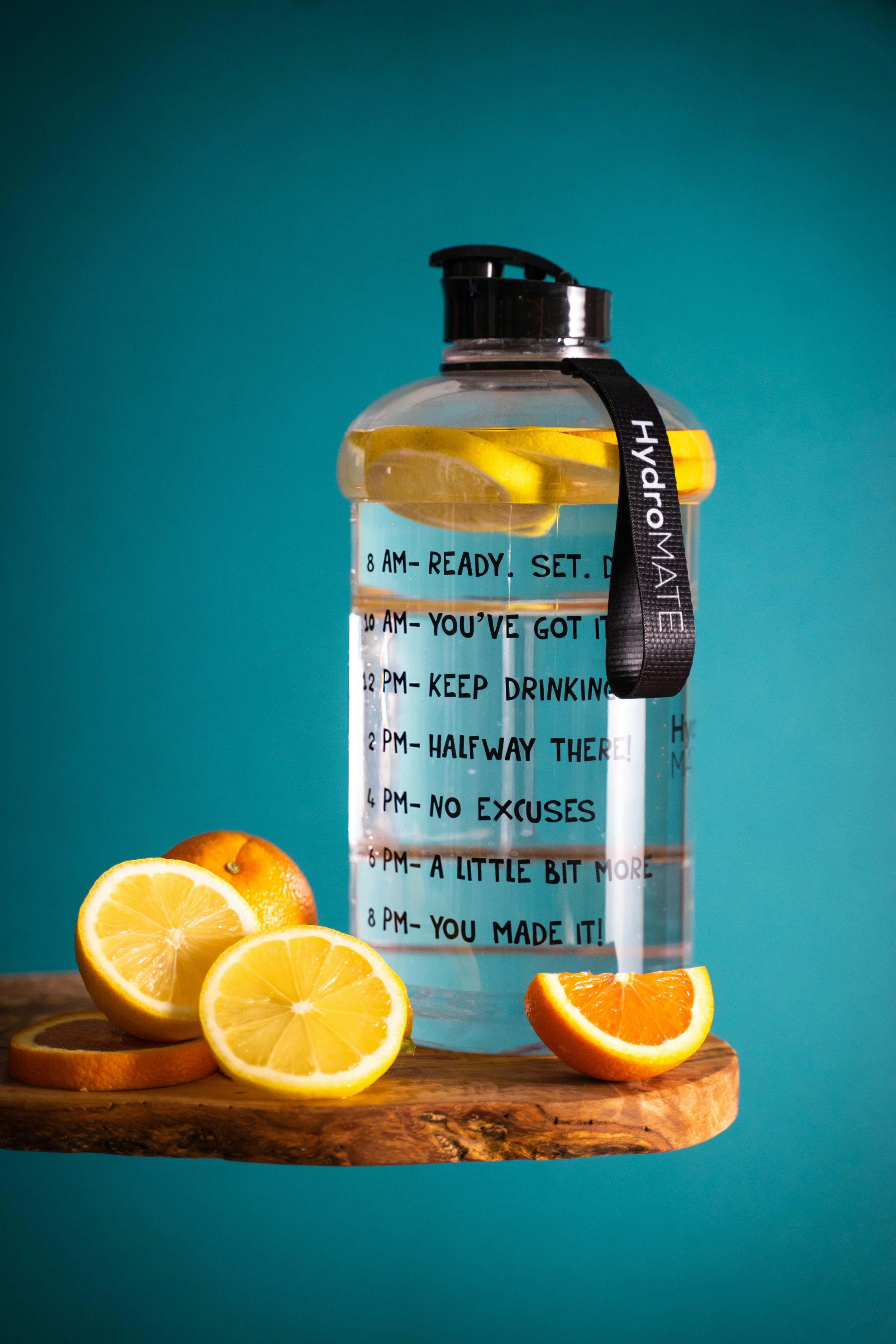 A transparent water jug with a black lid and strap, filled with water and lemon slices, placed on a wooden surface with lemon and orange slices beside it, against a teal background.