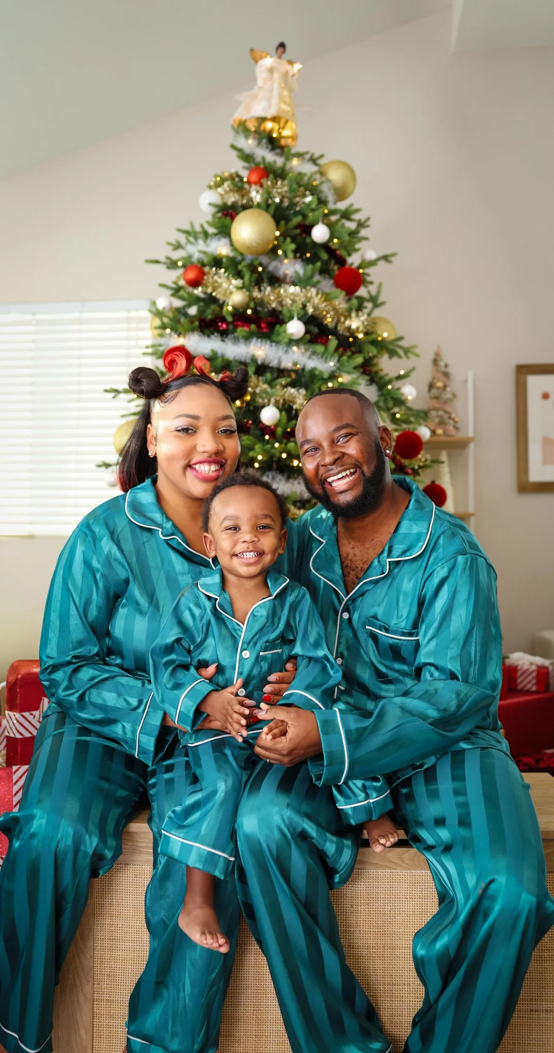 A happy family of three wearing matching teal pajamas sitting in front of a decorated Christmas tree, smiling for the camera.